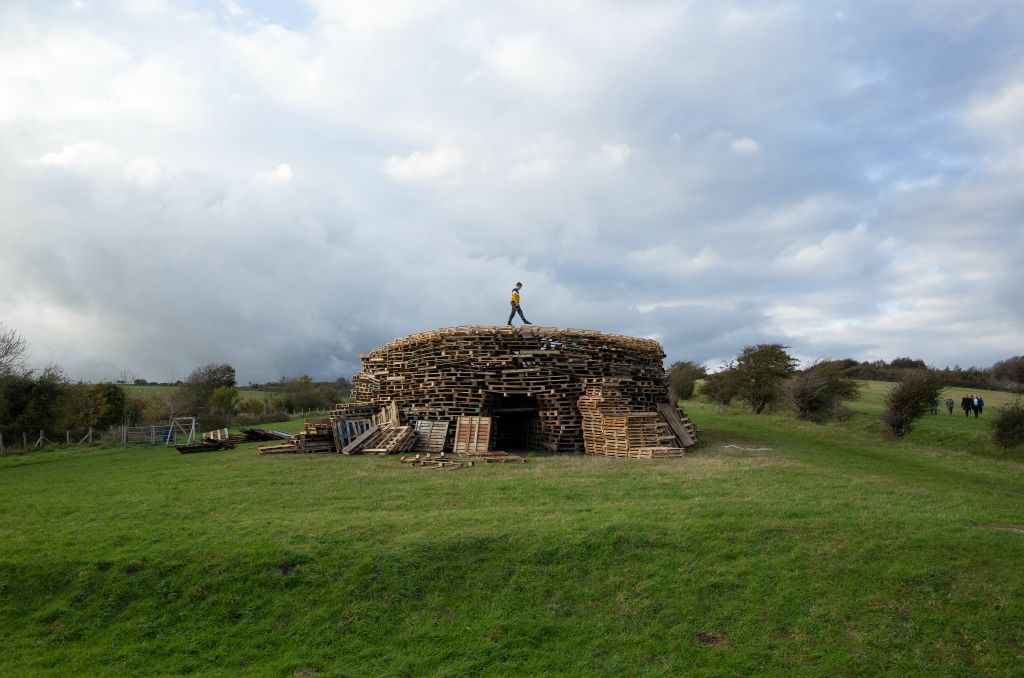 One of the fire site's at Lewes Fireworks Festival  midway through being built in a grass field. A boy is striding on the top of the wooden pallet structure and he is above a gaping entrance into the fire centre.