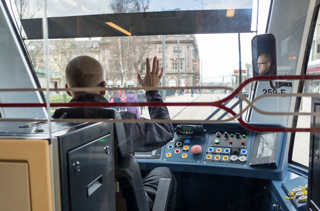A tram driver waves to another tram driver. He is photographed driving with his face reflected in the rear mirror.
