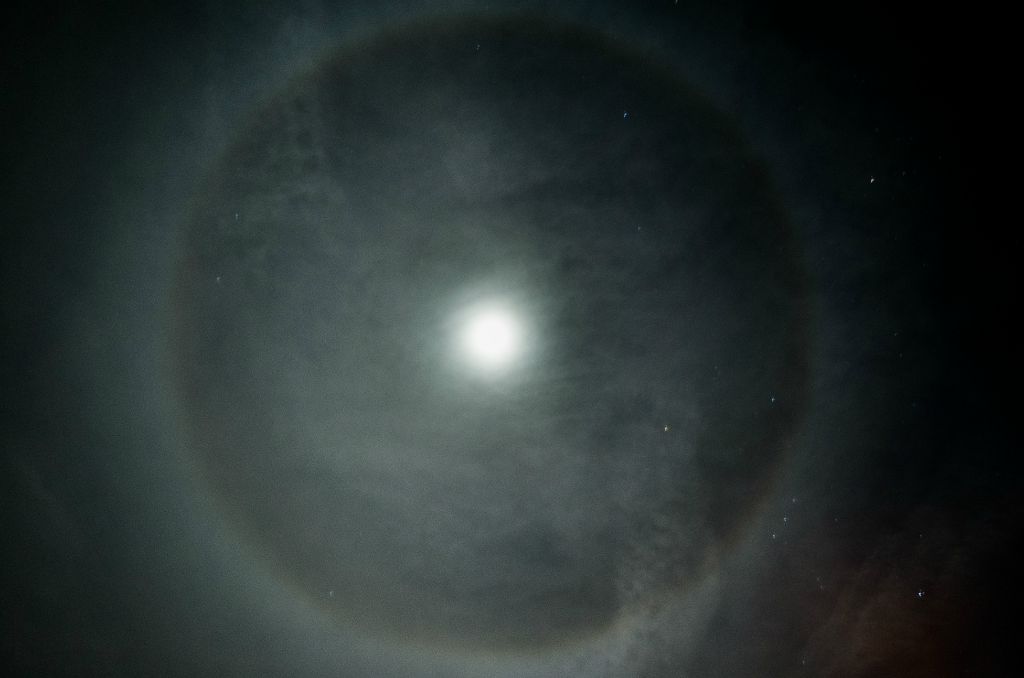 A nighttime photograph depicting a moon ring, an optical phenomenon caused by both refraction, or splitting of light, and reflection, or glints of light on tiny ice crystals.