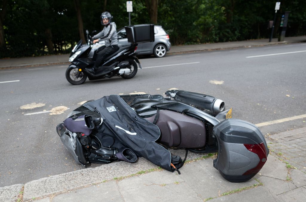 In the foreground of this landscape photo a new looking grey moped lies on the pavement after appearing to be knocked over. Another moped driver views the scene whilst driving past in the background.