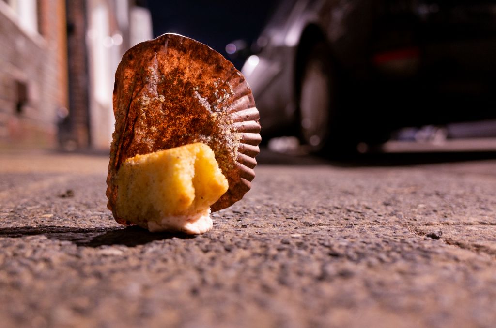 A discarded, half eaten cupcake lies on the pavement lit by street light at night.