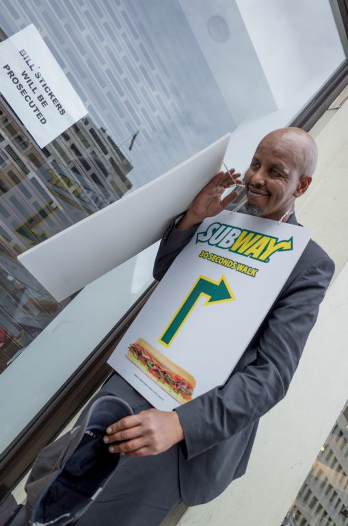 This portrait photograph depicts a brown gentleman who is struggling to put a double sided, wearable advertising board over his shoulders. The board is advertising Subway and has the strapline 30 seconds walk. The image is titlted at about 45 degrees which would be called a dutch angle in film production terms.