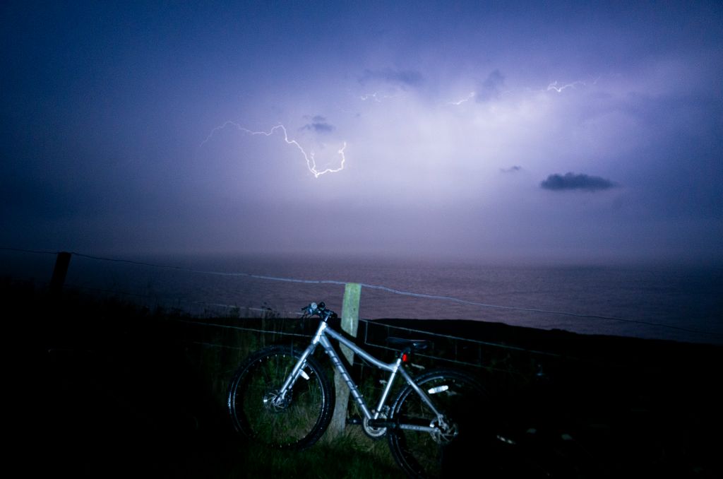 A seascape looking out towards a lightening storm with a bike in the foreground.