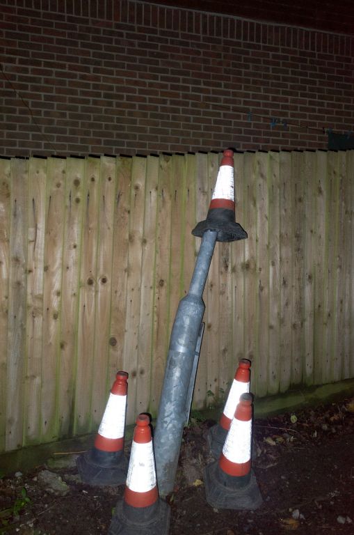 Five traffic cones surround a broken lamppost. Four surround the base and one is sat on top of the broken lamppost.