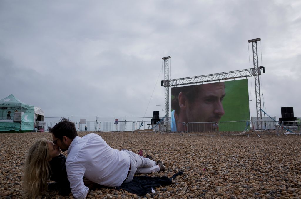 A couple kiss whilst reclined on a pebble surface. A large projected screen depicts Andy Murray's face in close up presumably playing tennis.