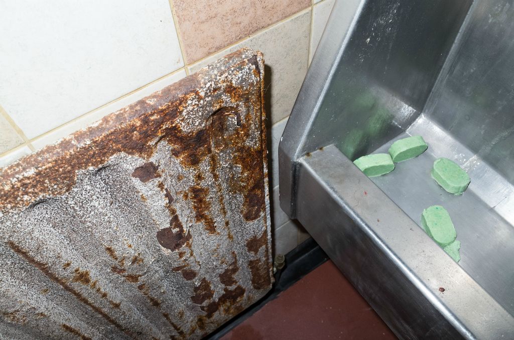 On the left of the landscape photograph is a very rusty radiator. On the right and far to close is the end of a stainless steel urinal trough with four green deodorising cakes in the trough.