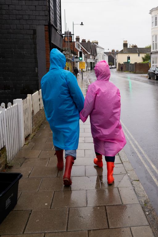 Rainy day walkers, one dressed in a pink waterproof, another dressed in a blue waterproof depicted form behind walk down a rainy street.
